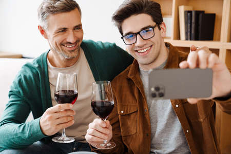 Happy gay couple hugging and drinking wine while taking selfie on cellphone at homeの写真素材