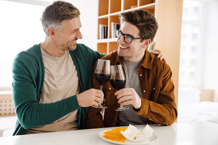 Happy caucasian gay couple hugging while drinking wine at homeの写真素材