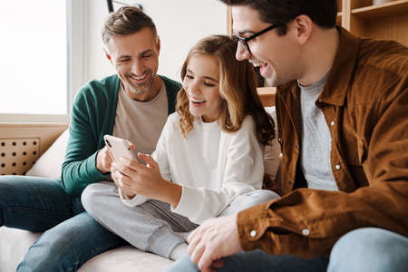 Happy gay family smiling and using mobile phone while sitting on couch at home
