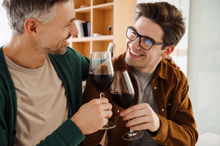 Happy caucasian gay couple hugging while drinking wine at homeの写真素材