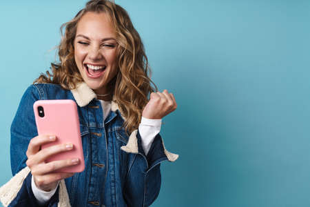 Blonde excited woman using mobile phone and making winner gesture isolated over blue wallの写真素材