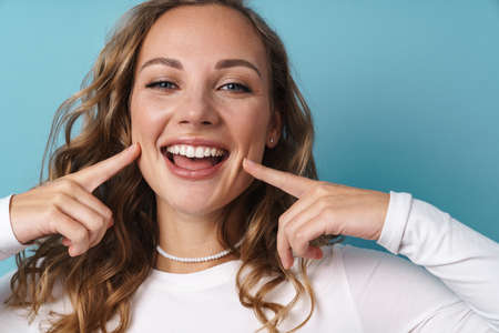 Blonde happy woman pointing fingers at her cheeks and smiling isolated over blue wallの写真素材