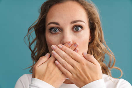 Blonde shocked woman covering her mouth and looking at camera isolated over blue wallの写真素材