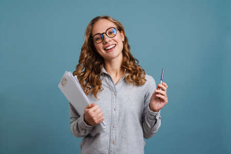 Happy girl in eyeglasses smiling while posing with papers isolated over blue backgroundの写真素材