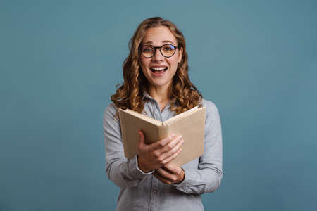 Smiling attractive young woman reading a book while standing isolated over blue backgroundの写真素材