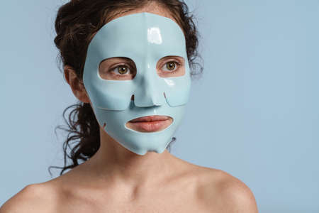 Beautiful half-naked nice girl posing with facial mask isolated over blue backgroundの写真素材
