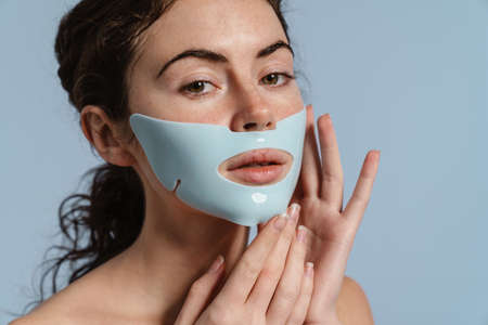 Beautiful half-naked nice girl posing with facial mask isolated over blue backgroundの写真素材