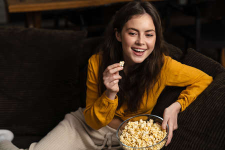 Young laughing woman eating popcorn while sitting on sofa at home