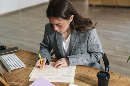 Young brunette woman writing down notes while working with computer in office