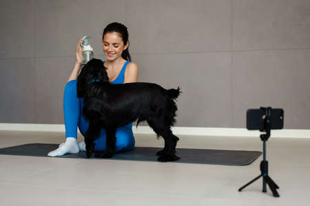 Happy young brunette woman blogger exercising with her dog on a fitness mat in front of camera...
