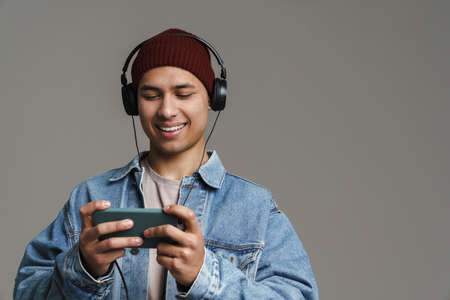 Joyful handsome guy listening music with headphones and cellphone isolated over grey background