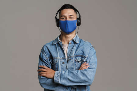 Brunette happy man in face mask listening music with headphones isolated over grey wallの写真素材