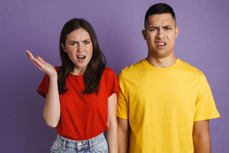 Brunette displeased couple frowning and gesturing at camera isolated over purple wall