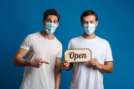 Pleased two guys in face masks holding and pointing finger at open sign board isolated over blue...