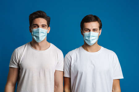 Pleased brunette two guys in face masks posing and looking at camera isolated over blue backgroundの写真素材