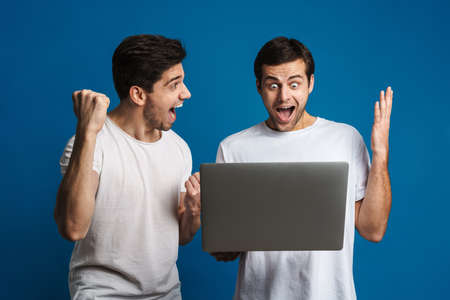 Excited two guys making winner gesture while posing with laptop isolated over blue backgroundの写真素材