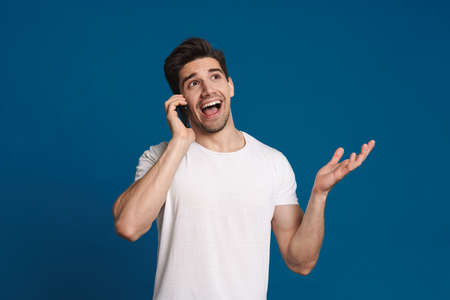 Happy unshaven guy smiling while talking on mobile phone isolated over blue backgroundの写真素材