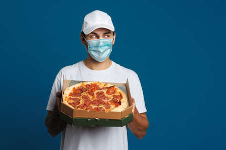 Happy caucasian delivery man in face mask posing with pizza isolated over blue backgroundの写真素材