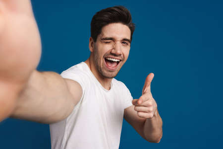 Happy guy winking and pointing finger at camera while taking selfie isolated over blue background