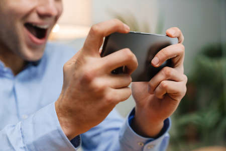 Happy young businessman using smartphone and celebrating success at workplace
