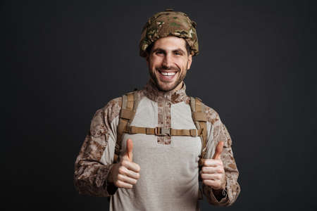Happy military man wearing helmet smiling and showing thumb up isolated over black backgroundの写真素材