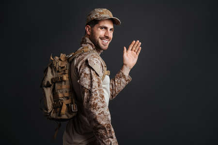 Smiling masculine military man waving hand while posing with backpack isolated over black background