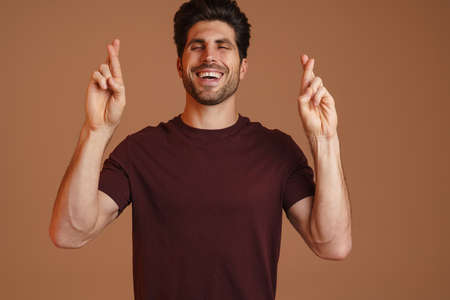 Joyful masculine man posing with fingers crossed for good luck isolated over beige background