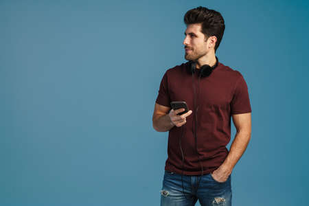 Pleased handsome man with headphones using mobile phone isolated over blue backgroundの写真素材