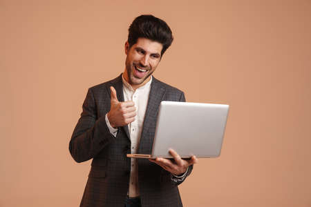 Joyful young man showing thumb up while working with laptop isolated over beige background