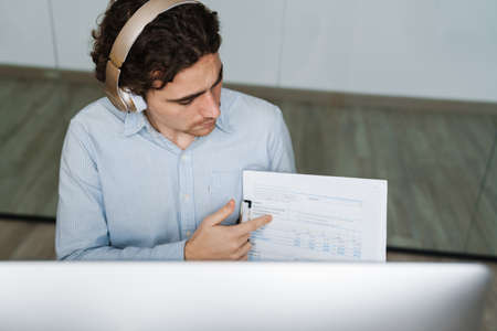 Caucasian young businessman in headphones showing paper documents at computer while working in officeの写真素材