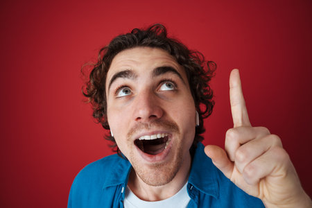 Excited handsome man in earphones pointing finger upward isolated over red backgroundの写真素材