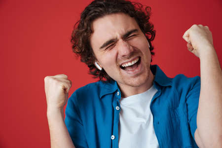 Excited handsome man in earphones making winner gesture isolated over red background