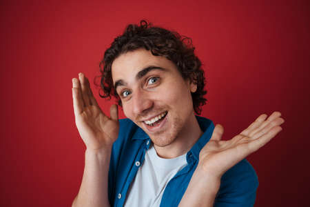 Amusing handsome curly man making fun on camera isolated over red backgroundの写真素材