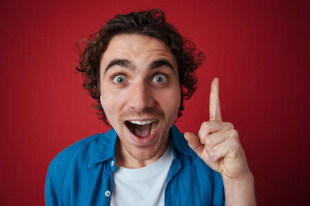 Excited handsome man in earphones pointing finger upward isolated over red background