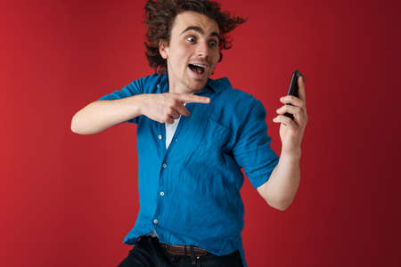 Excited man holding and pointing finger at his cellphone while jumping isolated over red background