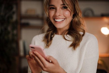 Close up of a smiling attractive woman using mobile phone while sitting in the modern officeの写真素材