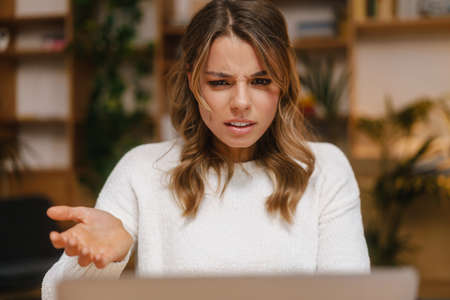 Frustrated confused young woman manager looking at laptop computer while sitting at the office desk,...