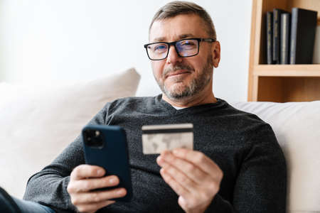 Pleased adult unshaven man in eyeglasses using cellphone and credit card at homeの写真素材