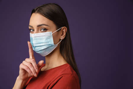 Close up of a young white woman wearing protective medical mask over violet wall background, showing...