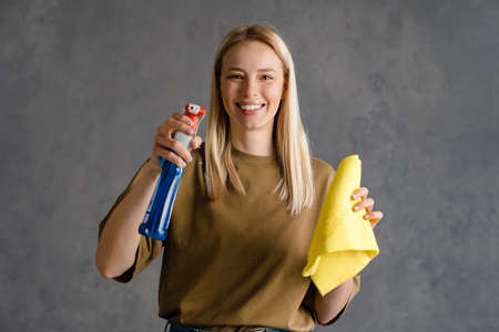Happy blonde woman maid holding bottle of detergent and a rag, spraying glassの写真素材