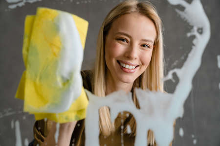 Happy european woman smiling and cleaning glass with rag isolated over grey wall