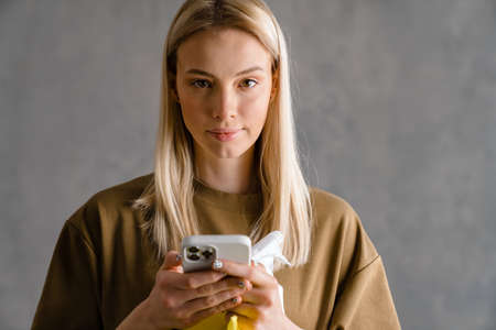 Young european woman using mobile phone and holding cleaning spray isolated over grey wallの写真素材