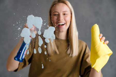 Happy blonde woman maid holding bottle of detergent and a rag, spraying glassの写真素材