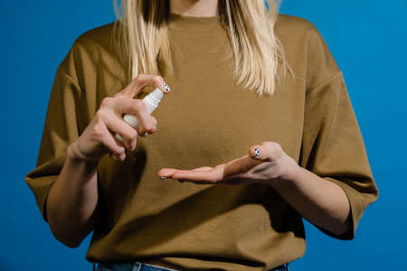 Blonde european woman using antiseptic spray for her hands isolated over blue wallの写真素材