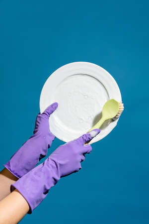 Closeup photo of hands wearing gloves washing plate with brush isolated over blue wallの写真素材