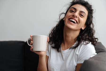 Happy brunette woman laughing and drinking coffee while sitting on sofa at homeの写真素材