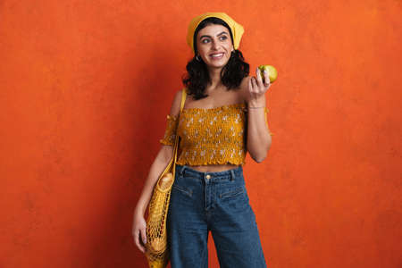 Joyful beautiful girl smiling while posing with bag and apple isolated over orange backgroundの写真素材