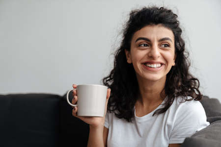Happy brunette woman smiling and drinking coffee while sitting on sofa at homeの写真素材