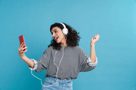 Portrait of a happy woman listening to music with headphones isolated on blue background, holding...