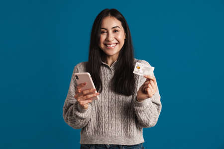 Happy beautiful asian girl posing with credit card and smartphone isolated over blue background